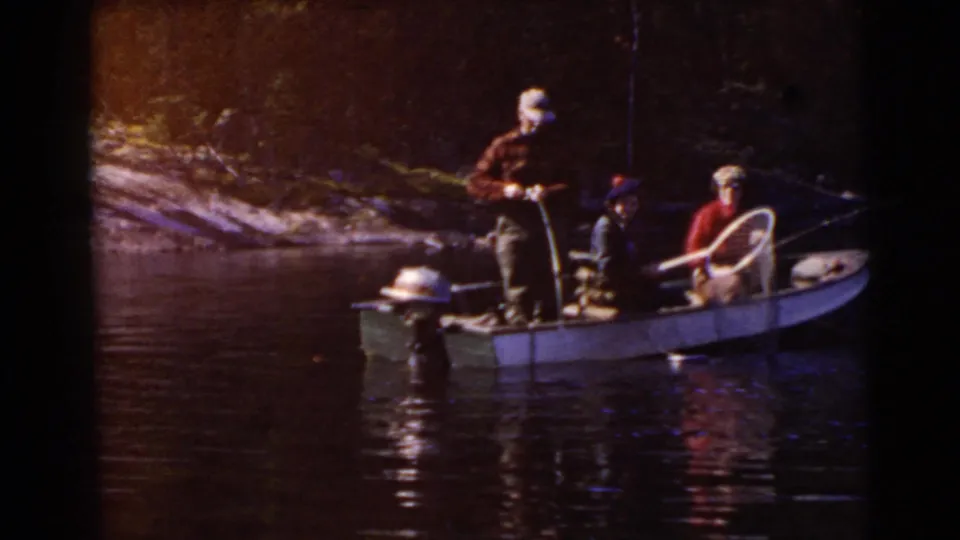 Wide view of adult men fishing from a small outboard motorboat near a wooded shoreline in the 1956 Miami home movie.