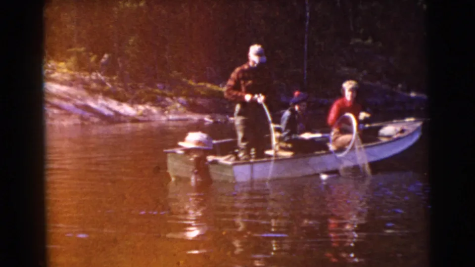 1956 Miami footage shows an adult standing near the outboard motor while another sits at the bow with fishing gear on a calm lake.