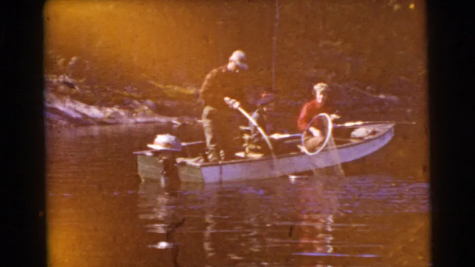 An 8mm 1956 Miami frame shows two adults in a small fishing boat, with warm exposure and a wooded shoreline in the background.