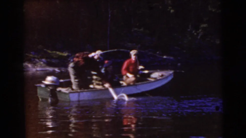 A darker 1956 Miami home-movie frame shows two adults in a small boat, with one leaning over gear as water reflects below.