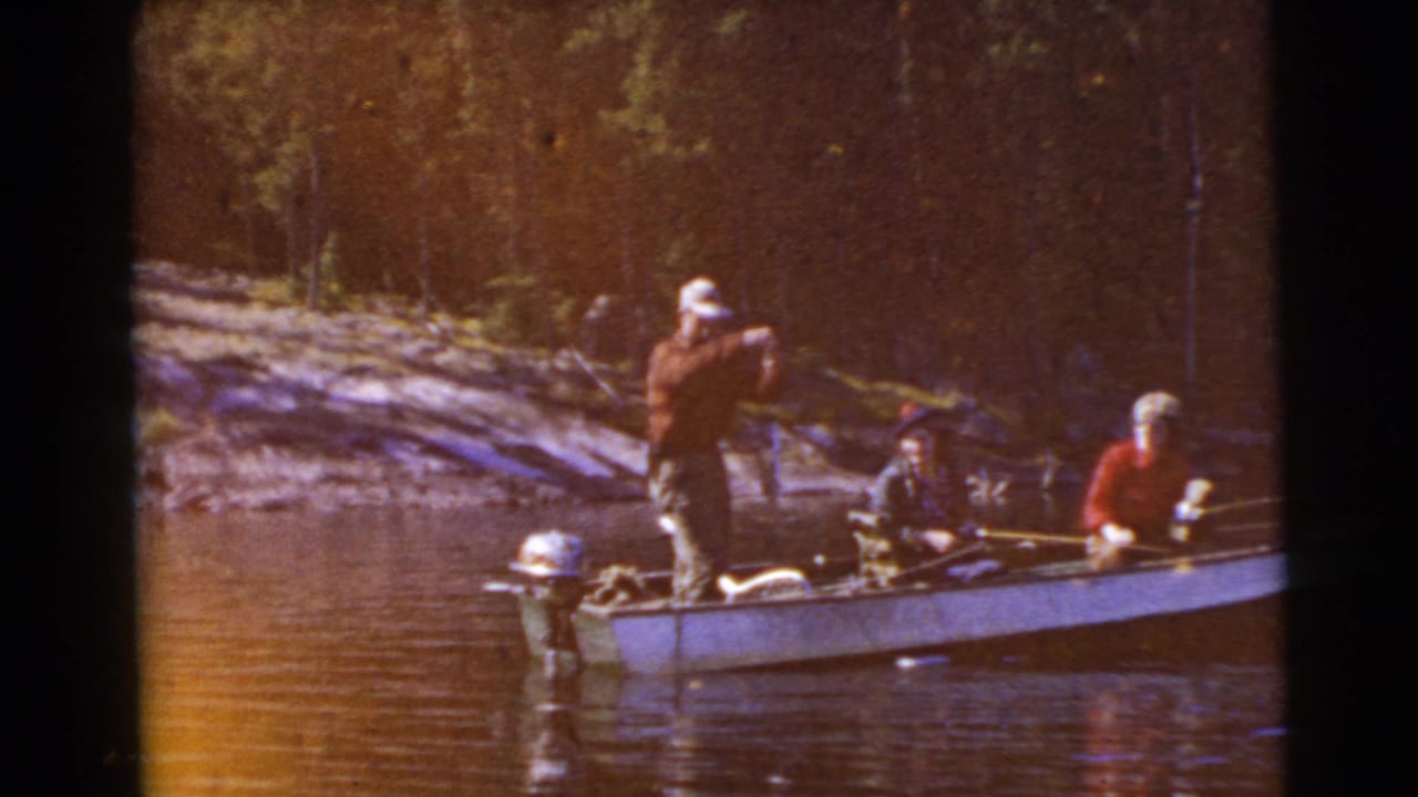 A standing person holds a fishing rod while two others sit in a small boat on the lake.