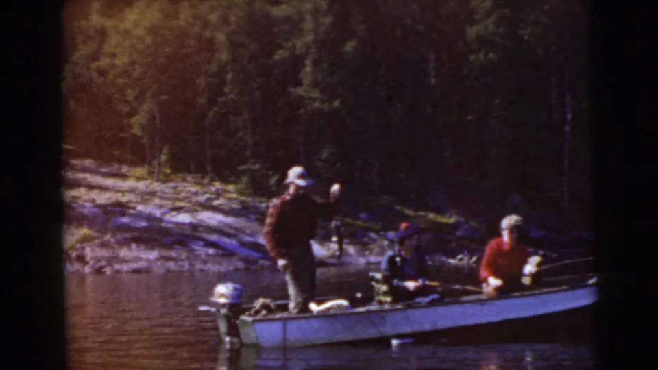 1956 8mm lake-fishing frame showing three people in a small boat close to a wooded shoreline, with fishing gear and an outboard motor visible.