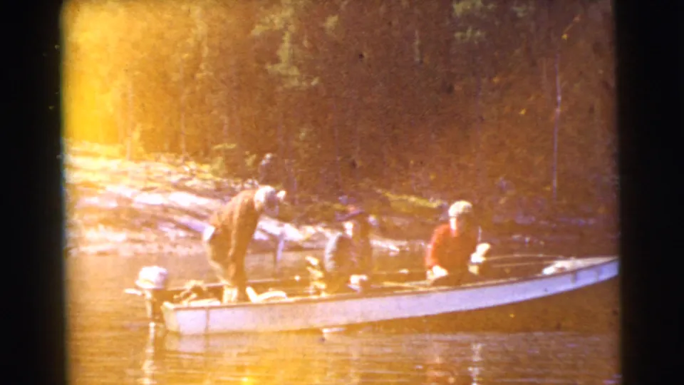 1956 Miami-area 8mm frame with warm exposure flare over three people fishing from a small motorboat on a lake beside a wooded shore.