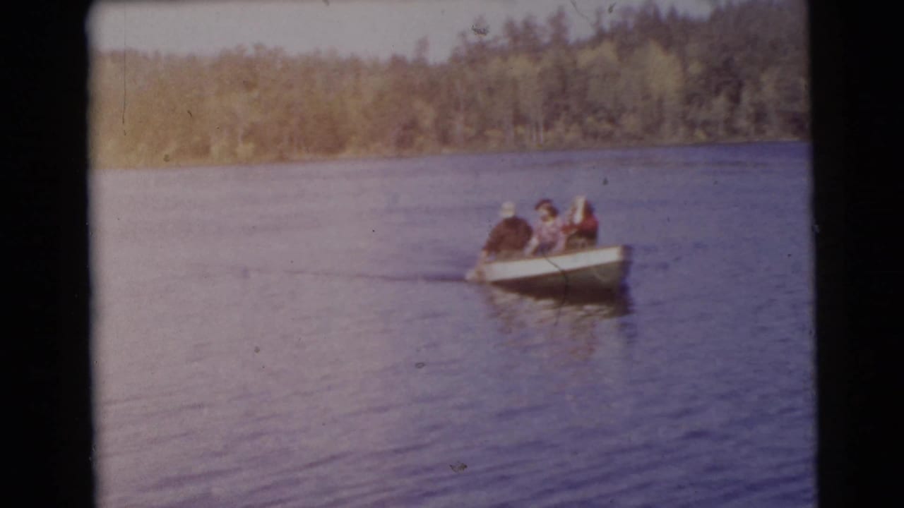 Three adults in a small rowboat on a calm lake with trees along the far shore.