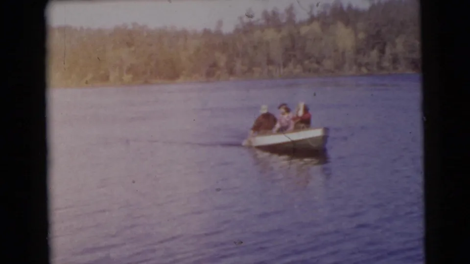 1956 Miami home-movie frame showing three adults in a small rowboat moving across a calm lake toward a wooded shoreline.