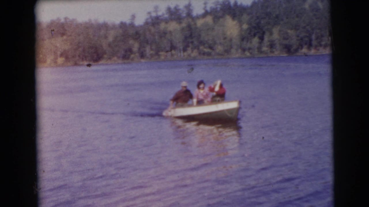 A small rowboat carrying three adults moves across open water near a tree-lined shore.