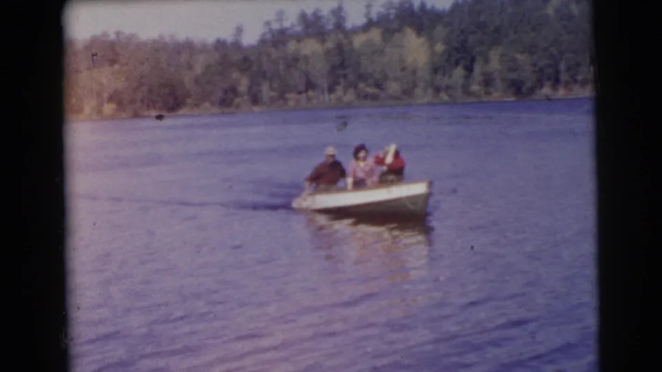 The rowboat remains mid-lake in this 1956 Miami 8mm frame, with three adults seated and the distant shoreline behind them.