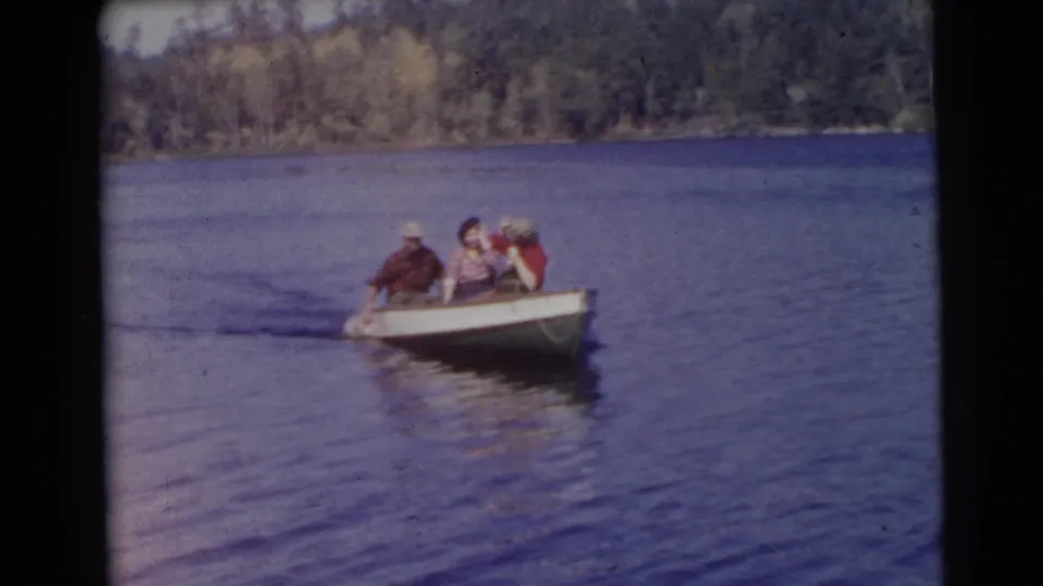 In the final 1956 Miami frame, three adults in a small rowboat approach across the lake, with rippled water and a wooded shore behind them.