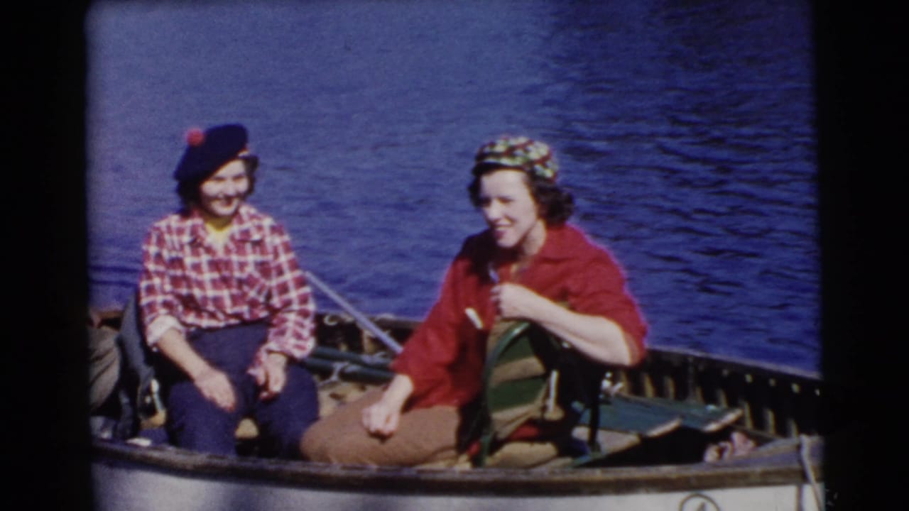 Two women seated in a small boat on blue water, one in plaid with a beret and one in a red jacket.