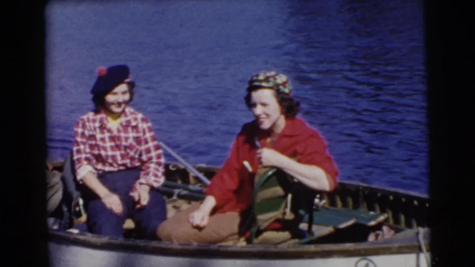 1956 Miami home-movie frame of two women sitting in a small open boat on a sunny lake, wearing casual midcentury clothing.