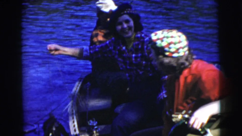 1956 Miami home-movie frame showing a smiling woman in plaid seated in a boat beside a companion wearing a bright patterned cap.