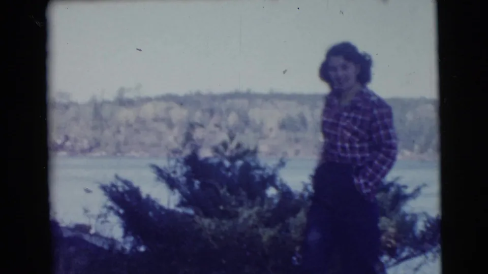 1956 Miami home-movie frame of a woman in a plaid shirt standing near shoreline shrubs with water and a distant bank behind her.