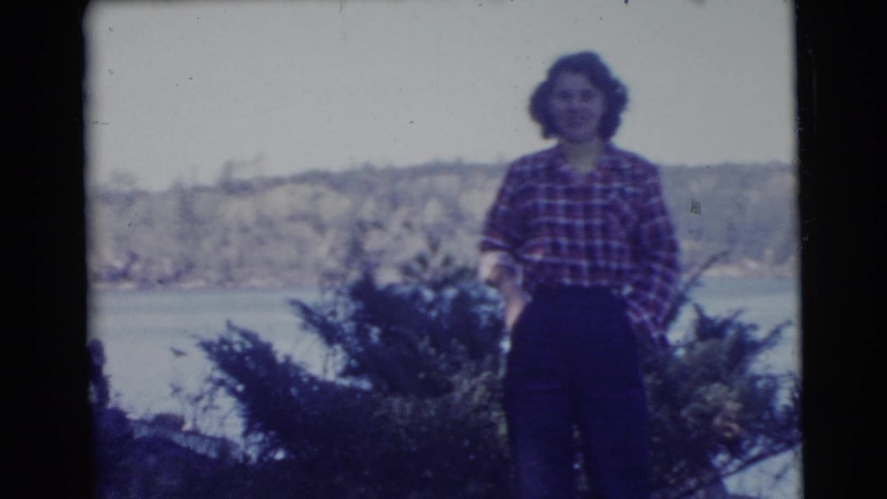 Woman in a red plaid shirt standing near brush with water and trees behind her.