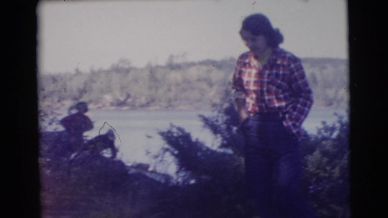 Woman in plaid shirt standing in profile near shrubs with water in the background.