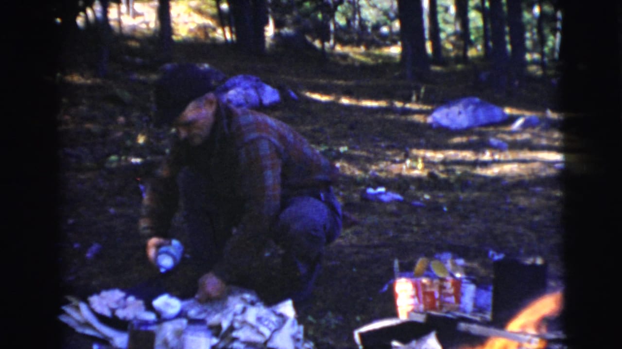 Adult crouches near camp food and a small fire in a wooded area.