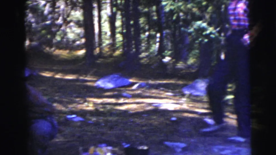 An adult in a checkered shirt and dark trousers appears in a wooded campsite, with rocks and scattered camp items visible.