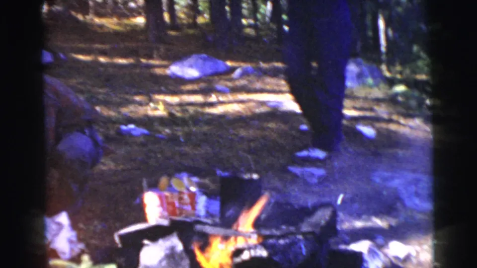 Two adults share a wooded campsite scene, with one crouched near supplies and another standing beside a small fire.