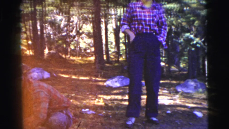 An adult in a checkered shirt stands among trees in the 1956 wooded camping scene, with another person partly visible near the ground.