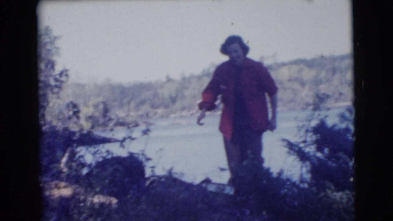 A woman in a red jacket walks through wooded ground beside a lake in supported 1956 Miami home-movie footage.