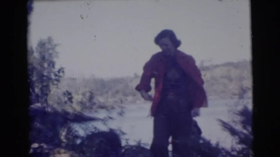 The woman continues across uneven wooded ground with a lake and distant trees in the background.