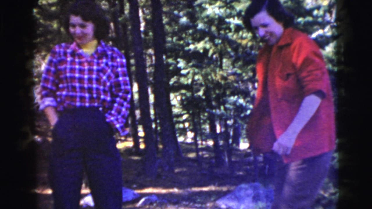 Two women standing among trees, one in plaid and one in a red jacket.