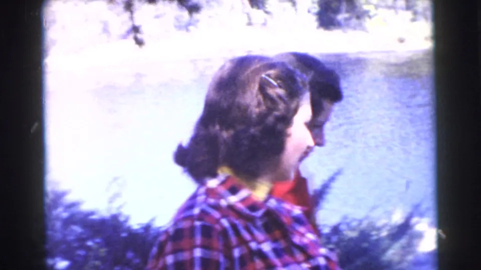 Adult woman in a red and blue plaid shirt stands near bright lakeside water, with a companion partly visible behind her.