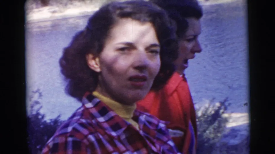 Close 8mm frame of a woman in a plaid shirt by the water, with another adult woman in a red jacket behind her.