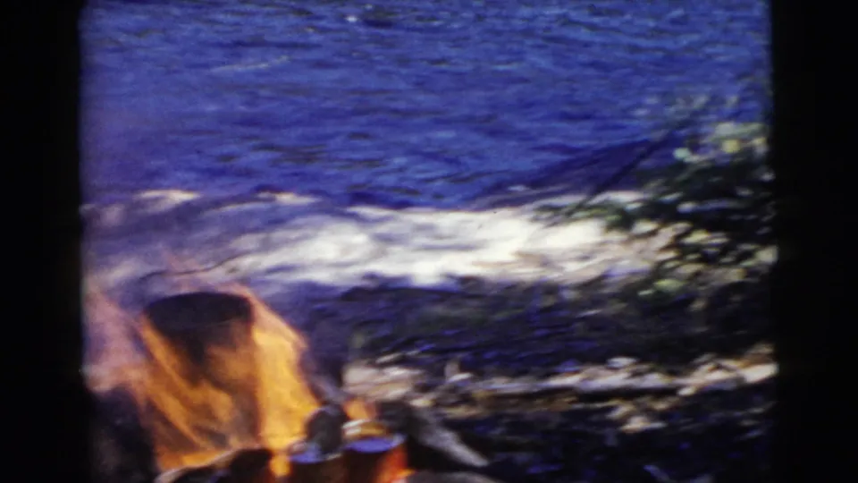 Campfire flames beside a blue lake in 1956 Miami-area home-movie footage, with brush and shoreline visible.