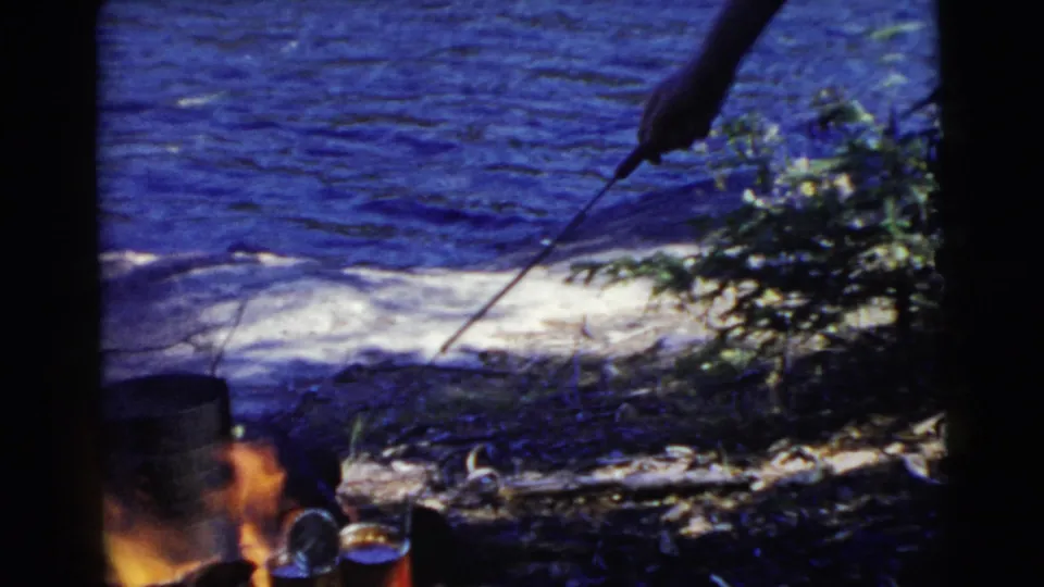 A person holds a roasting stick over a campfire with cooking cans near a Miami-area lake in 1956 home-movie footage.