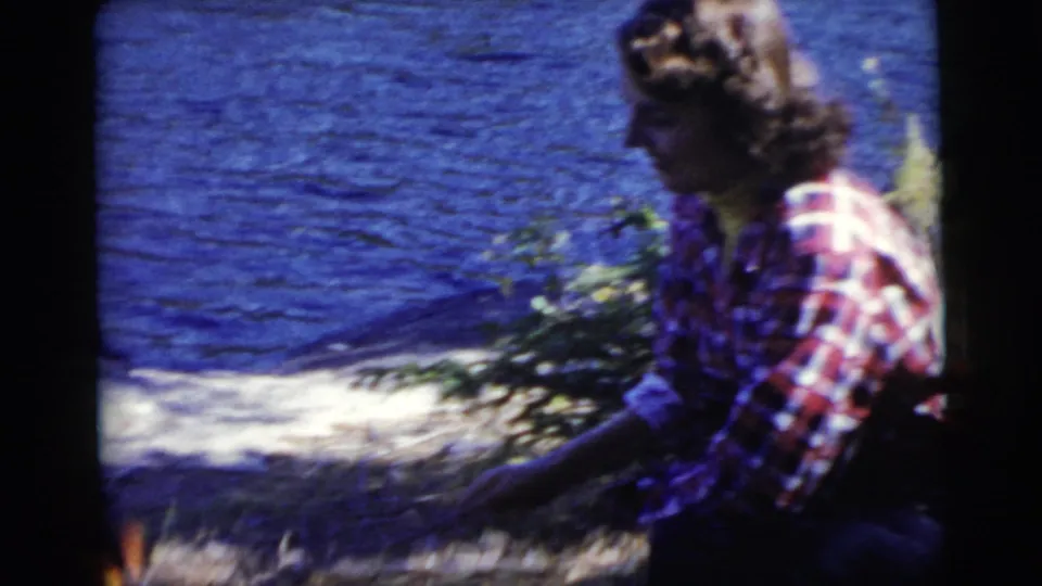 A person in a plaid shirt sits near a lakeside campfire in 1956 Miami-area 8mm home-movie footage.