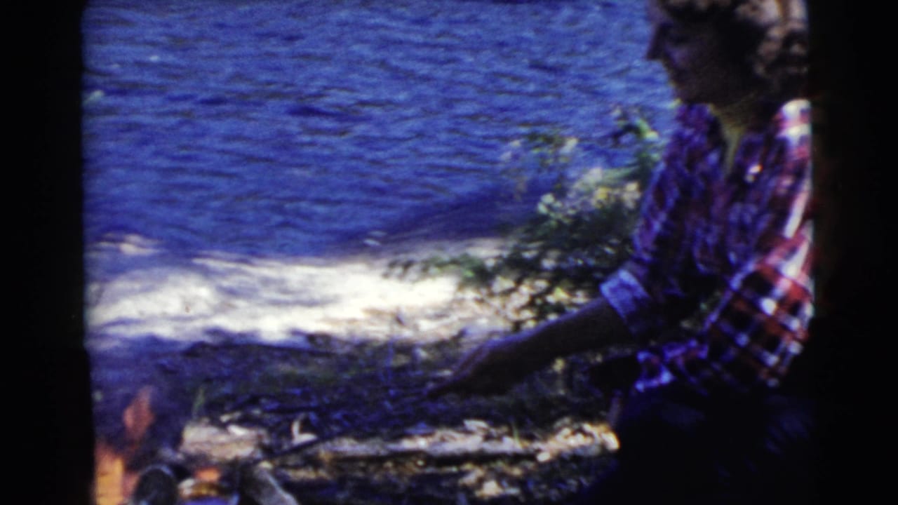 Woman in a plaid shirt crouching beside a campfire near blue lake water.