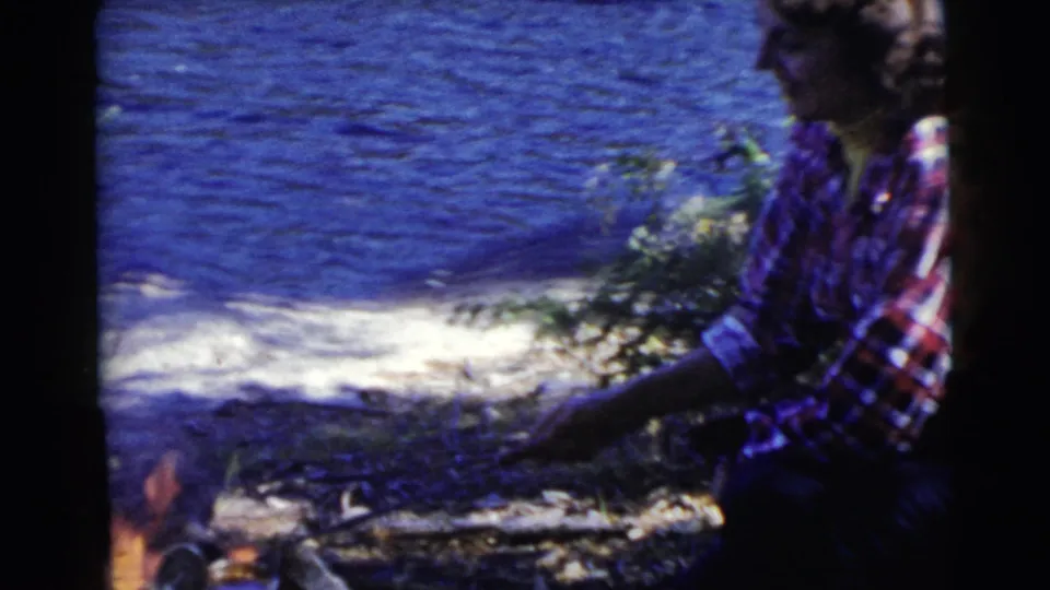 1956 Miami home-movie frame of a woman in a plaid flannel shirt crouched by a small campfire near a lake shore.