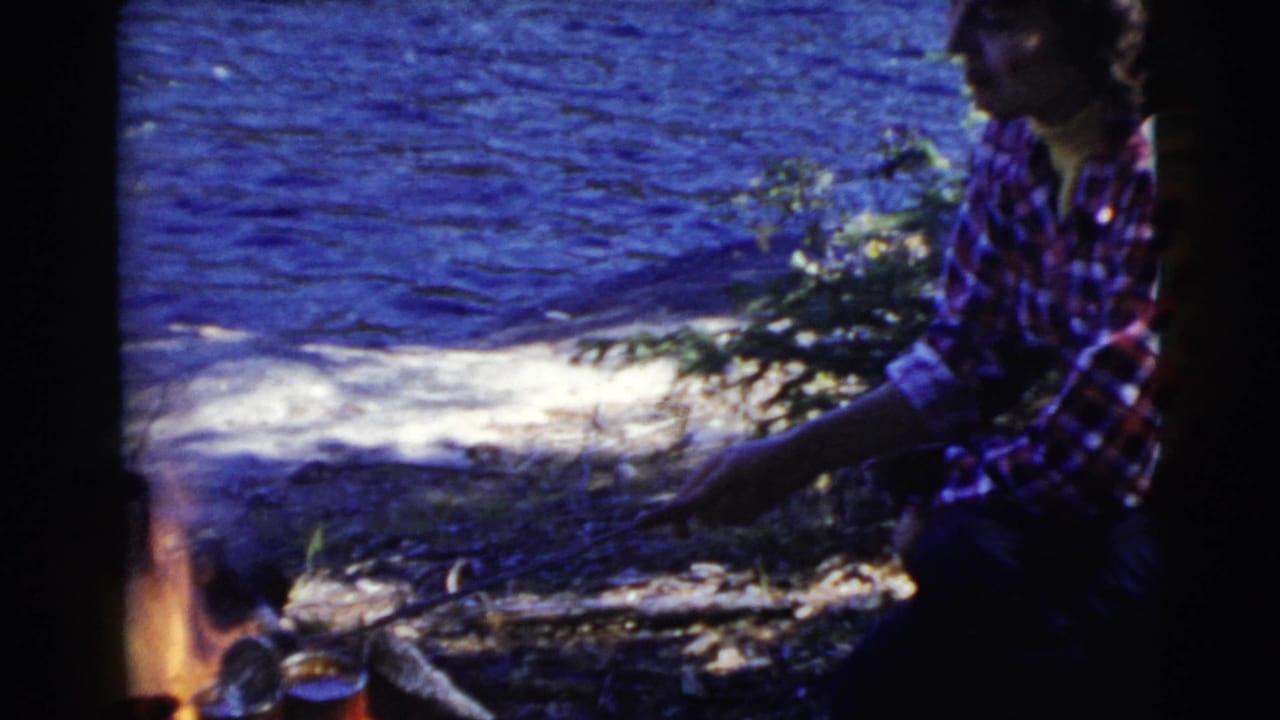 The woman remains beside the fire at the lake edge, with a small pot or pan near the flames in the 8mm frame.