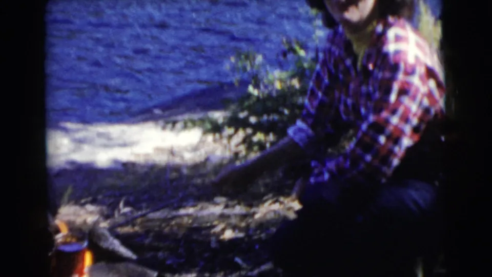 A soft, handheld 8mm frame shows the plaid-shirted woman beside the shoreline fire with lake water behind her.