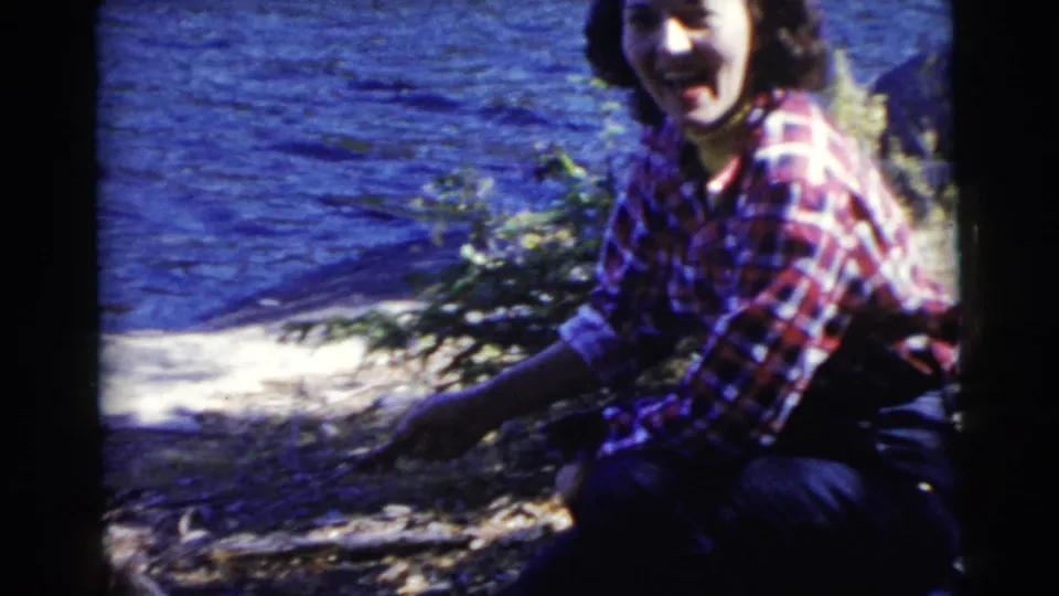 In a clearer 1956 Miami frame, the woman smiles toward the camera while crouching by the lakeside campfire.