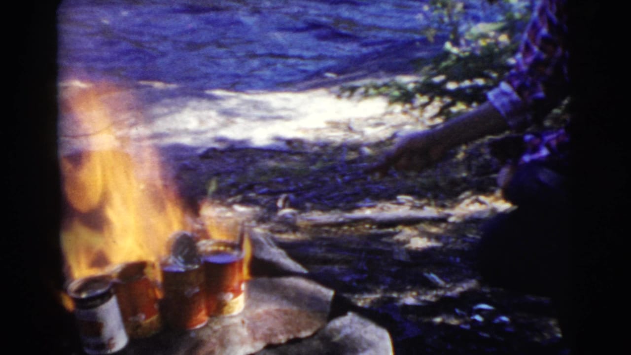 Campfire heating several cans on rocks near lake water and a crouching woman partly visible.