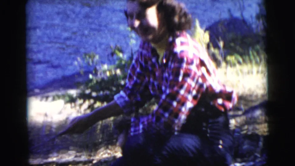 In this 1956 Miami home-movie frame, the woman crouches near the lakeshore in a plaid shirt, slightly blurred by motion and film softness.