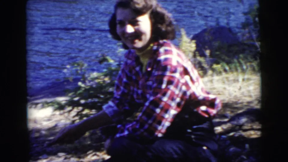 A smiling woman crouches near the lakeside campfire area in a 1956 Miami home-movie frame, with water and trees behind her.