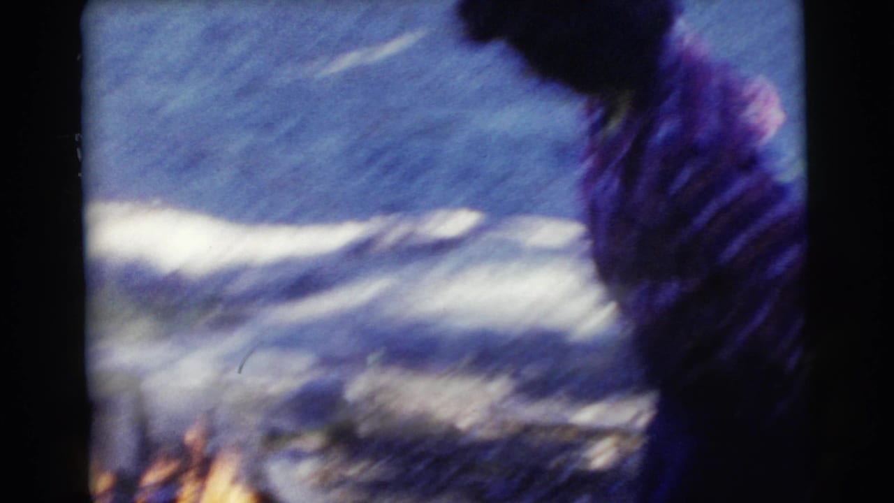 Woman in a plaid shirt near water with a small campfire at the lower left.