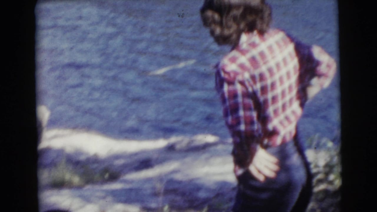 Woman seen from behind in a plaid shirt beside water and rocks.