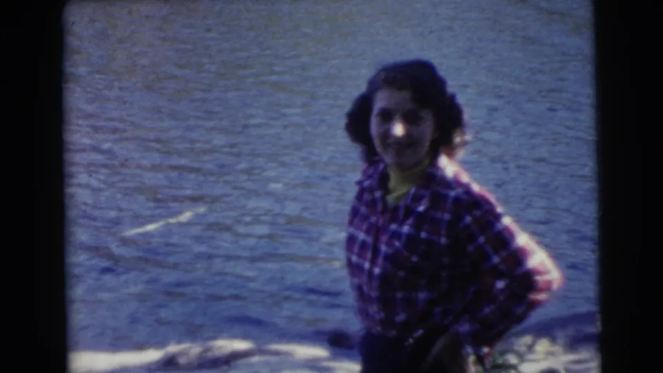 1956 Miami-area home-movie still of a woman in a red plaid shirt smiling toward the camera beside water.
