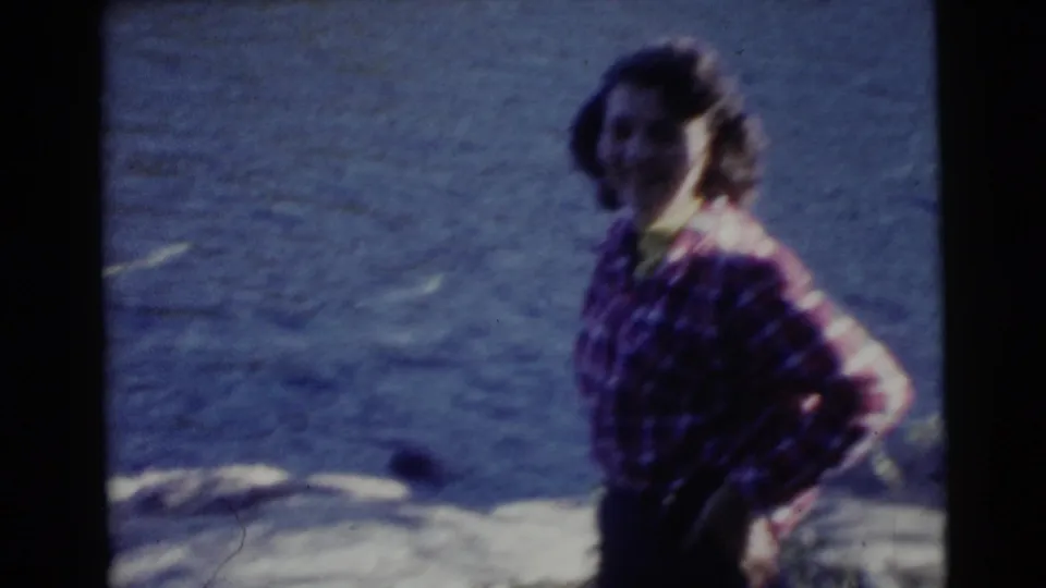 Soft 8mm frame from 1956 Miami showing a woman in a plaid shirt posing near shoreline water.