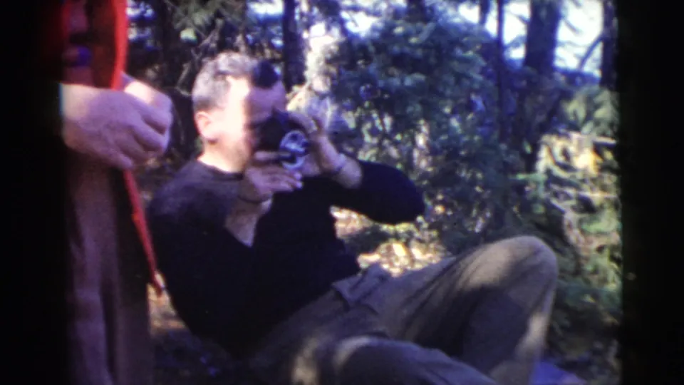 A man in a dark sweater lies on the ground and drinks from a vessel as a person in a red jacket stands nearby in the woods.