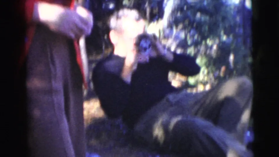A reclining man drinks in a wooded 1956 Miami home-movie frame while a person in red stands partly cropped at left.