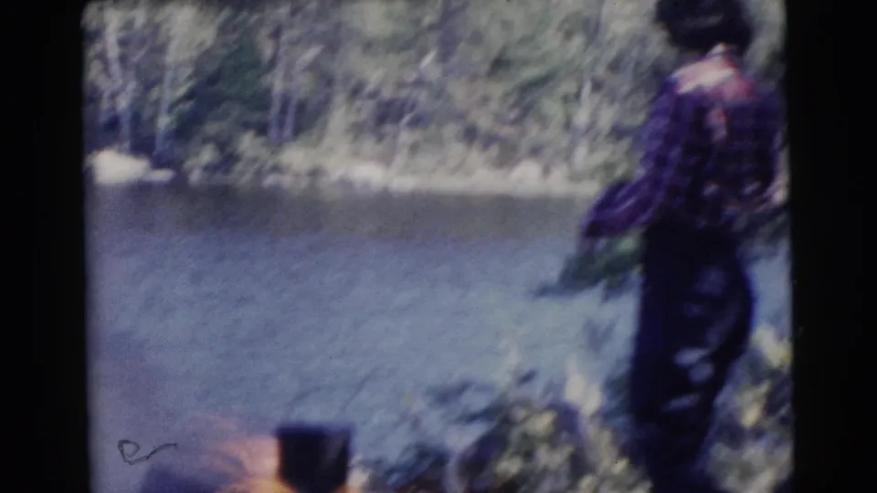 A person in a plaid shirt stands near a calm riverbank with trees and shoreline vegetation in the background.