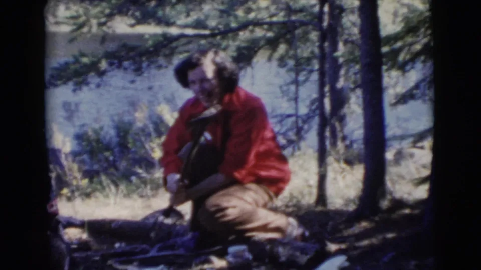 A woman in a red shirt kneels by branches or logs in a shaded wooded area beside water in 1956 Miami home-movie footage.