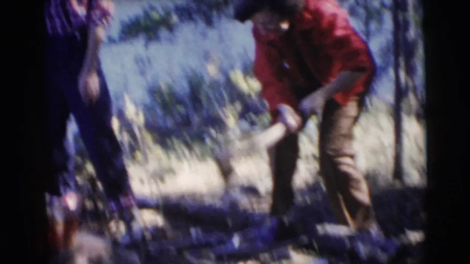 A woman in a red shirt bends over wood with an axe as another adult stands beside her in the wooded riverside setting.
