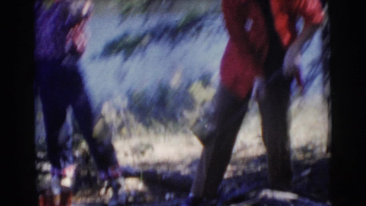 Two women stand outdoors near a lakeside campsite, with one partly blurred in a red jacket.