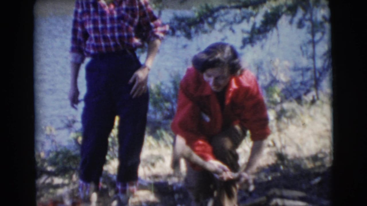 A woman in a red jacket bends over firewood while another woman in a plaid shirt stands nearby.