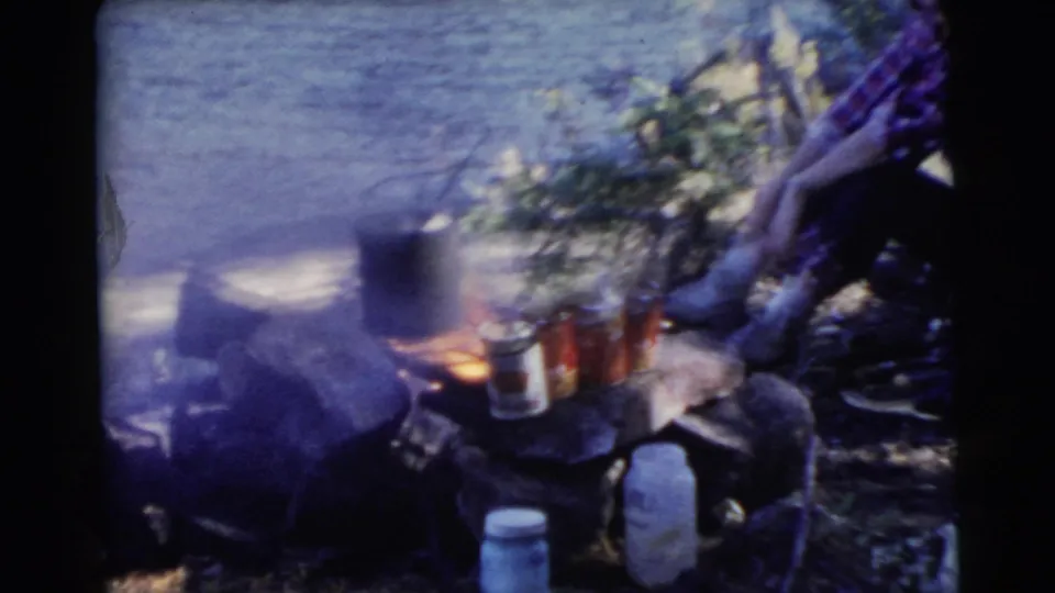 A lakeside campfire burns beside cans and simple supplies, with a seated figure partly visible at the edge of the 8mm frame.
